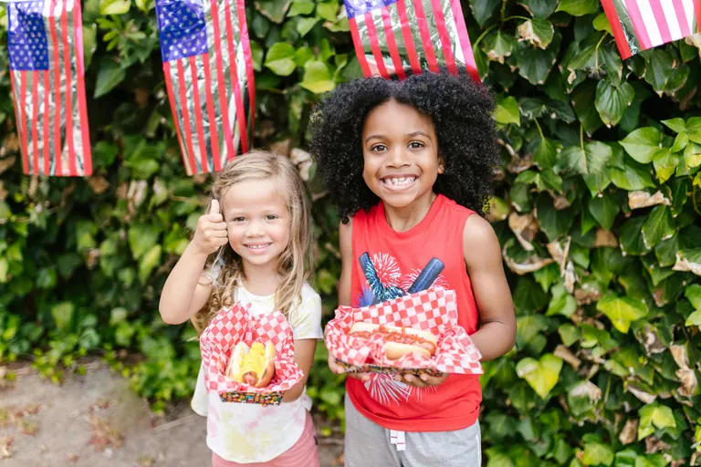 Two smiling children holding hot dogs at an outdoor cookout, American flags hanging behind them